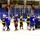A post-game handshake line is taking place on an indoor ice hockey rink, with two teams — one in black and gold jerseys and one in blue jerseys — meeting at center ice. A referee in a black-and-white striped shirt is visible among the players. The arena has blue bleacher seating with a modest crowd of spectators watching from the stands.
