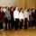 A group of approximately 13 children and young teenagers are standing in a line on a wooden stage or platform, appearing to sing in a choir or perform at a school event. They are dressed in a mix of casual and semi-formal attire, with several wearing school-style uniforms including plaid skirts. The setting appears to be an indoor school hall or community room with gold curtains and a television monitor visible in the background.