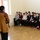 Two adults, one male and one female, stand facing a group of approximately 20 seated students in what appears to be a classroom or meeting room. The students, who appear to be middle-school aged, are seated in chairs arranged along the wall and are attentively facing the two standing adults. The setting suggests a formal presentation, orientation, or guest speaker event.