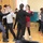 A group of young people are participating in a dance class or social dance activity in what appears to be a community hall or school room. Several pairs are dancing together in a ballroom or folk dance style, with adults appearing to guide or instruct some of the younger participants. The setting is informal, with a wooden floor, yellow curtains, and minimal furniture.