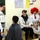 A group of children and young teenagers appear to be performing a skit or theatrical activity in a classroom setting. Several students are wearing costumes, including a bright red curly wig and a blonde wig, suggesting a role-play or drama exercise. The setting is clearly a school classroom with educational posters and a chalkboard visible on the walls.