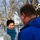 Two individuals are engaged in a conversation outdoors during winter. A woman with short dark hair wearing a gray knit sweater and teal ruffled scarf faces the camera, while a man in a blue jacket with glasses has his back to the camera. The setting appears to be near a fenced playground or park area with snow-covered ground and bare trees.