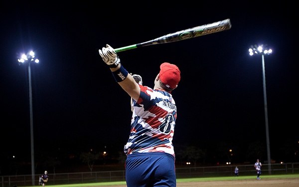 A baseball/softball player with a prosthetic leg is captured mid-swing or follow-through at a night game under stadium lights. The player is wearing a red, white, and blue patterned jersey and a red cap, viewed from behind. The scene takes place on a dirt infield with other players visible in the background.