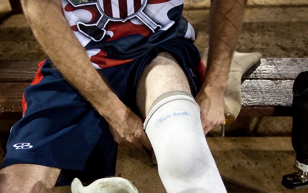 A person wearing a USA athletic uniform is seated on a wooden bench, managing prosthetic limbs. The individual appears to be an amputee athlete, handling a residual limb covered in a white prosthetic sock labeled 'Soft-Sock' while prosthetic legs are visible nearby. The setting appears to be a locker room or dugout environment.