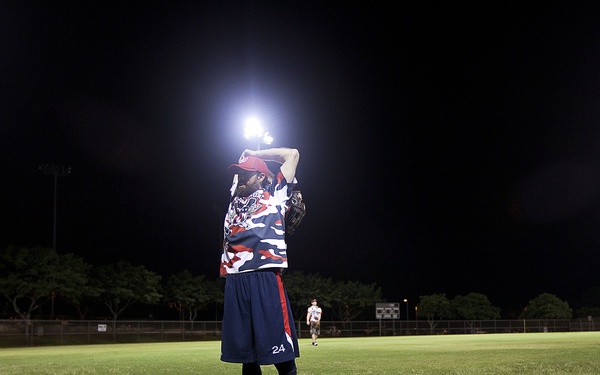 A nighttime baseball or softball game scene showing three players in matching red, white, and blue camouflage uniforms on a grass field. Two players are lying on the ground while a third stands upright wearing number 24, appearing to react to a play. The field is illuminated by stadium lights against a dark sky.
