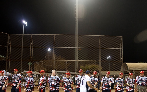 A nighttime softball or baseball event featuring a team of players wearing matching red, white, and blue patriotic uniforms lined up along the baseline of a dirt field. Several players are visibly wearing prosthetic limbs, suggesting this is a wounded warrior or adaptive sports event. A person in a white jersey walks along the line, possibly during introductions or a pre-game ceremony.