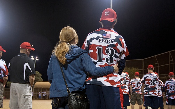 A nighttime softball game featuring players wearing red, white, and blue camouflage uniforms. The central focus is on player #13 'Clark,' who has a prosthetic leg, standing arm-in-arm with a woman in a blue hoodie. The scene appears to be a military or veterans' charity softball event, with multiple uniformed players lined up along the field.