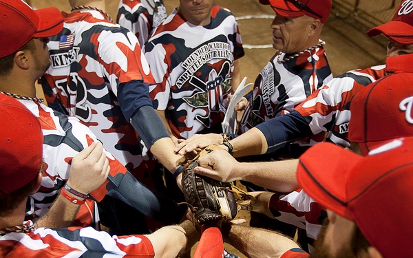 A group of players wearing red camouflage uniforms and red baseball caps are gathered in a team huddle, with hands stacked together at the center. The jerseys display text referencing 'Wounded Warrior Amputee Softball' team, indicating this is a charity or military-affiliated softball event. Several players are also wearing Washington Nationals 'W' caps, suggesting involvement with the MLB organization.