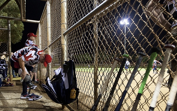 A nighttime softball or baseball game is in progress, photographed from inside a dugout. Multiple bats are leaning against the chain-link fence in the foreground, while players in red and white camouflage uniforms occupy the dugout area. Field lights illuminate the playing field visible in the background.