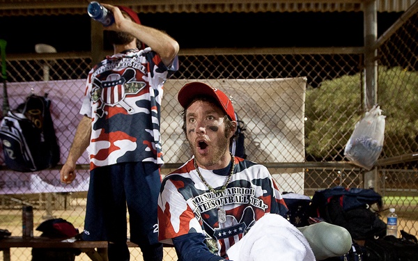 Two men wearing camouflage-patterned uniforms reading 'Wounded Warrior Softball Team' are in a dugout area. The seated man in the foreground wears a red cap, eye black, a gold chain, and has a prosthetic leg visible, holding a white towel. The standing man in the background is pouring water from a bottle over his head.