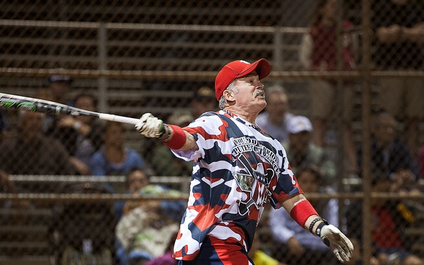 A middle-aged male softball player has just completed a full swing at bat during a night game, with the bat visible in follow-through motion. He is wearing a patriotic red, white, and blue camouflage-style uniform with matching accessories, including a decorated leg guard or prosthetic on his right leg. Spectators are visible seated in bleachers behind a chain-link fence in the background.