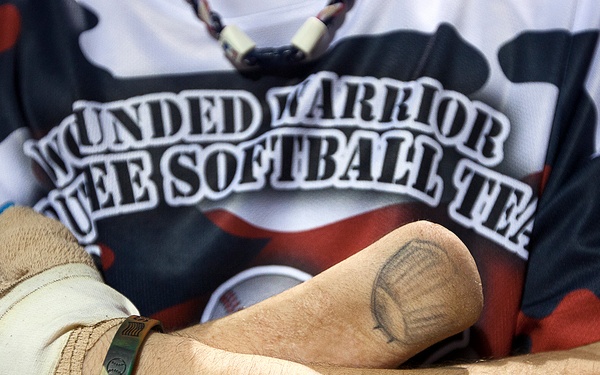 A close-up photograph of a person's crossed arms wearing a 'Wounded Warrior' softball team jersey in a camouflage-style red, white, and blue pattern. The individual has visible tattoos on their forearm and wrist area, wears a gold/brass bracelet inscribed with 'WWA' or similar text, and has a bandaged or prosthetic hand. The image highlights symbols associated with military service and veteran athletic programs.