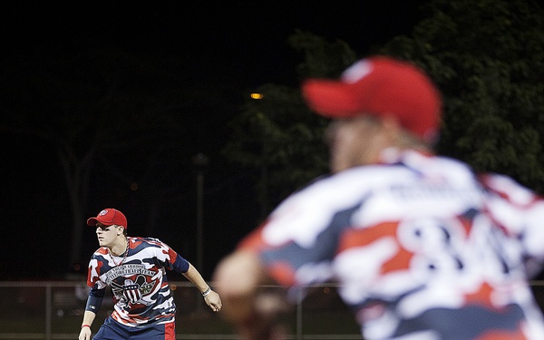 A nighttime softball or baseball game featuring players in matching red, white, and blue camouflage-patterned uniforms. Two players are visible on the infield dirt, with one standing near second base in a ready defensive stance and another in the foreground near first base. The uniforms appear to bear a 'Wounded Warrior' softball team logo.