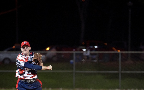 A nighttime recreational softball game is in progress on a dirt infield. The primary player in the foreground, wearing jersey number 12 and a red 'W' (Washington Nationals) cap, is in the motion of throwing the ball after fielding it. A second teammate is visible in the background near the outfield grass.