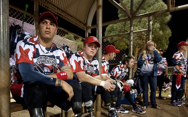 A group of athletes wearing matching red, white, and blue camouflage softball uniforms sit in a dugout during a nighttime game. Several players visibly have prosthetic legs, indicating this is a wounded warrior or adaptive sports event. Spectators and additional team members stand in the background.