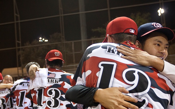 A nighttime baseball or softball game scene showing players in red, white, and navy camouflage-patterned jerseys engaged in a post-game handshake or congratulatory line. The foreground features two players hugging, with jersey number 12 visible on one player's back. Multiple teammates are visible in the background participating in the same tradition.
