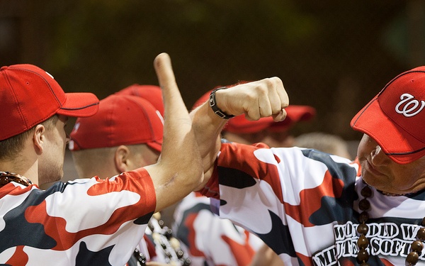 A group of players wearing matching red, white, and black camouflage uniforms from the 'Wounded Warrior Amputee Softball Team' are celebrating together, appearing to fist-bump or cheer. The players are wearing red Washington Nationals-style caps with a 'W' logo and uniforms featuring an American flag patch and individual names/numbers. The atmosphere appears energetic and celebratory.