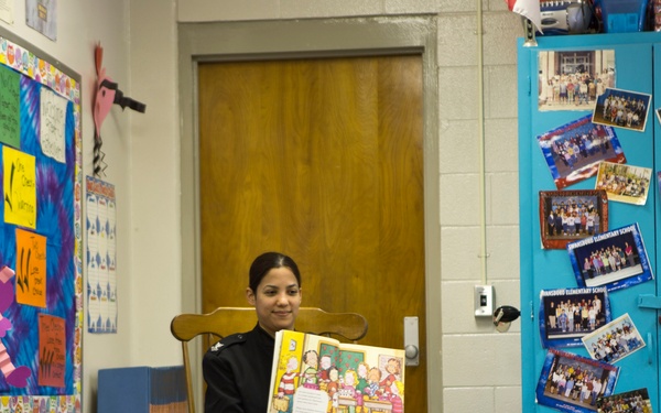 A woman in a black uniform is seated in a wooden chair reading a illustrated children's book aloud to a group of elementary school students in a classroom. The children are seated on the floor facing her attentively. The classroom is decorated with colorful bulletin boards, student artwork, and school photos.