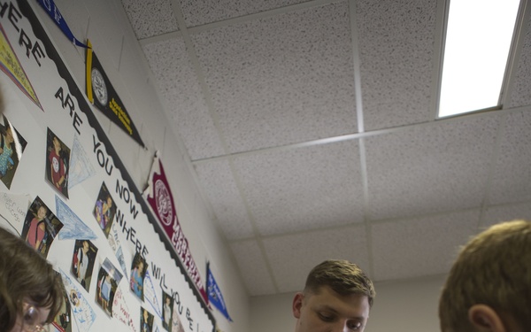 A young man in a dark olive/green military-style sweater is stirring contents in a red pot placed on a portable electric hot plate, apparently demonstrating cooking to children in a classroom setting. The classroom walls are decorated with bulletin boards, maps, pennants, and photographs. Two children are visible on either side of him observing the activity.