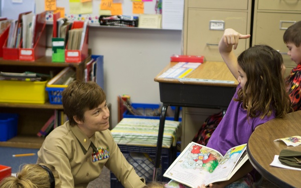 A military officer in tan/khaki uniform with service ribbons is reading a picture book to a group of young children in an elementary school classroom. The children are gathered around the officer, with one girl in a purple shirt raising her hand and holding the colorful picture book. The setting appears to be a classroom reading activity or special visit.