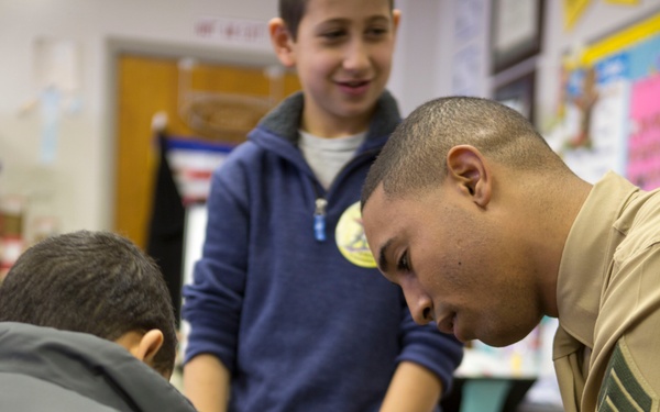 A person in a tan military-style uniform is kneeling down to interact with a young child in a gray jacket, inside what appears to be a classroom setting. A second child in a blue hoodie stands nearby holding a brown teddy bear and smiling. The scene suggests a community outreach or school visit by a service member.