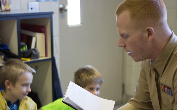 A U.S. Marine in service uniform is reading aloud from a book to a group of young children in what appears to be an elementary school classroom. The Marine holds the open book and leans toward the children, who are seated on the floor listening attentively. The setting includes classroom storage cubbies filled with backpacks and books in the background.