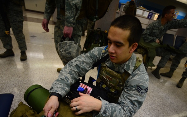 A U.S. Air Force service member in ABU (Airman Battle Uniform) is organizing or packing a large olive-drab military rucksack in what appears to be a terminal or staging area. The airman is wearing a tactical vest with a U.S. flag patch and has a Senior Airman rank insignia visible. Multiple other uniformed personnel are visible in the background, suggesting a unit deployment or movement operation.