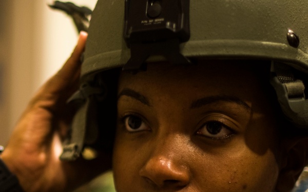 A female soldier in U.S. military uniform is adjusting or fitting a tactical combat helmet on her head. She is wearing digital camouflage (ACU pattern) and body armor, with a name tape partially visible at the bottom reading 'JACK...' The image is a close-up portrait shot with shallow depth of field, emphasizing her focused expression.