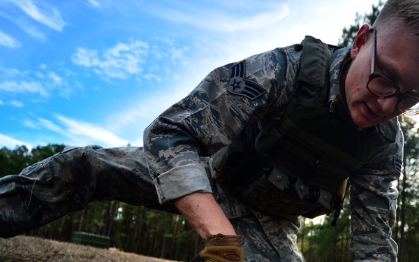 A U.S. Air Force enlisted member in ABU (Airman Battle Uniform) is performing a low-crawl or ground movement exercise in an outdoor field training environment. The airman is wearing tactical gloves, glasses, and a plate carrier/vest, and appears to be navigating through dry grass terrain. The setting suggests a field training exercise (FTX) or combat skills training course.