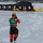 A winter emergency response training exercise is taking place on a frozen lake or body of water. Multiple responders in yellow and red cold-water survival suits are working around a large black inflatable structure, while a foreground figure in a green vest and red suit appears to be documenting or observing the exercise. Residential buildings are visible in the background across the frozen water.