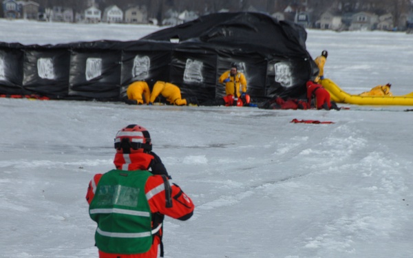 A winter emergency response training exercise is taking place on a frozen lake or body of water. Multiple responders in yellow and red cold-water survival suits are working around a large black inflatable structure, while a foreground figure in a green vest and red suit appears to be documenting or observing the exercise. Residential buildings are visible in the background across the frozen water.