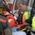 Emergency responders are loading an injured patient onto a stretcher near an ambulance. The patient is wearing a Coast Guard survival suit and orange helmet, and has been fitted with a cervical collar, suggesting a potential spinal precaution. EMS personnel and a firefighter are working together to manage the patient's care.