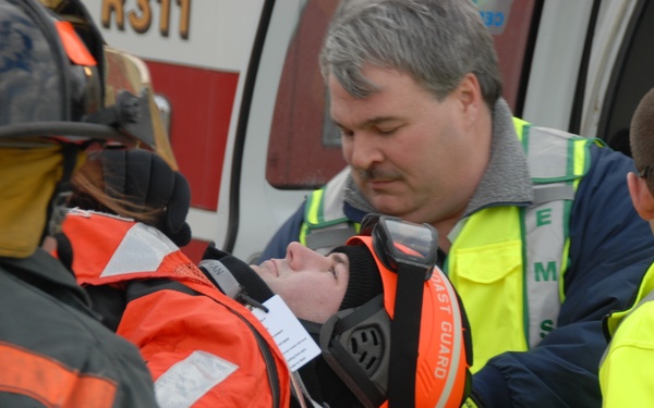 Emergency responders are loading an injured patient onto a stretcher near an ambulance. The patient is wearing a Coast Guard survival suit and orange helmet, and has been fitted with a cervical collar, suggesting a potential spinal precaution. EMS personnel and a firefighter are working together to manage the patient's care.