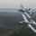 Two U.S. Air Force A-10 Thunderbolt II (Warthog) aircraft are flying in close formation over a heavily forested, hilly landscape under overcast skies. Both aircraft are painted in standard gray livery, with one featuring distinctive nose art on its forward fuselage. The image appears to be taken from a third aircraft flying alongside or above the formation.