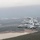 A formation of four A-10 Thunderbolt II (Warthog) attack aircraft flying in a staggered echelon formation over a forested landscape with a visible body of water below. The aircraft display markings consistent with the United States Air Force, with tail codes reading 'FS' visible on multiple aircraft. The lead aircraft features distinctive nose art and unit insignia markings.