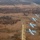 An aerial photograph shows four A-10 Thunderbolt II (Warthog) attack aircraft flying in a diagonal echelon formation over a rural, sparsely wooded landscape. The aircraft are painted in standard USAF gray livery and appear to be on a training or demonstration flight. Below them, a dirt road or runway strip is visible cutting through the terrain, with what appears to be a military installation or range facility in the upper right.