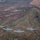 Four A-10 Thunderbolt II (Warthog) ground-attack aircraft are flying in a loose formation over a hilly, sparsely vegetated landscape. The aircraft are photographed from above and slightly behind, likely from a fifth aircraft or aerial platform. The terrain below appears to be late autumn or early winter, with bare deciduous trees and a prominent conical hill visible.