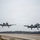 Two A-10 Thunderbolt II (Warthog) aircraft are captured simultaneously taking off or landing on a military airstrip under overcast skies. Both aircraft are in standard gray camouflage paint schemes with landing gear deployed. The image appears to be taken from ground level near the runway threshold, with a treeline and sparse infrastructure visible in the background.