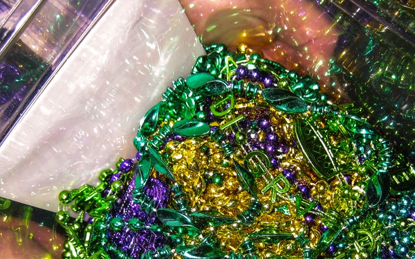 A clear plastic container is filled with a large collection of Mardi Gras beads and novelty items in the traditional colors of purple, green, and gold. The beads vary in size and style, including round beads, leaf-shaped beads, and decorative charms or pendants. A white plastic bag or packaging material is partially visible in the upper left corner of the container.