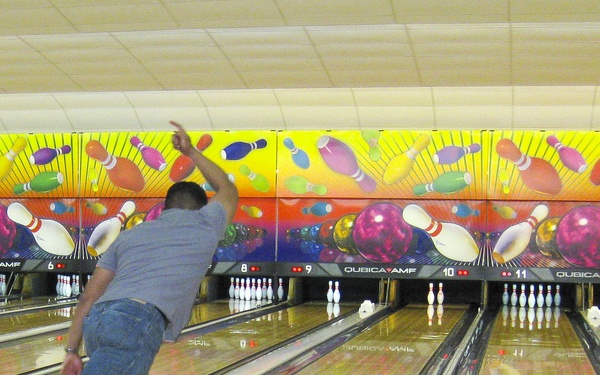 A man is captured mid-release while bowling at a QubicaAMF bowling alley. He is positioned on what appears to be lane 7 or 8, having just released a dark bowling ball that is visible on the lane. Multiple lanes with full pin sets are visible in the background, decorated with colorful bowling-themed murals along the back wall.