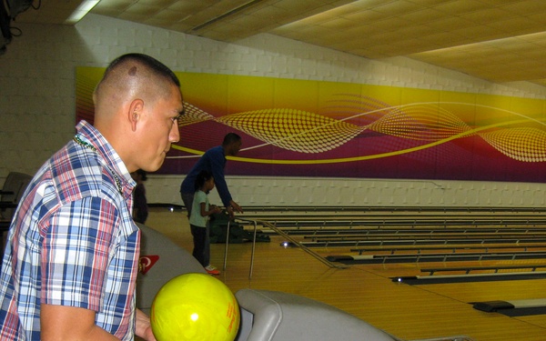 A man in a plaid shirt is holding a yellow bowling ball at a bowling alley, preparing to bowl. Multiple lanes are visible in the background along with other bowlers. The facility features a colorful purple and yellow wave mural along the back wall.