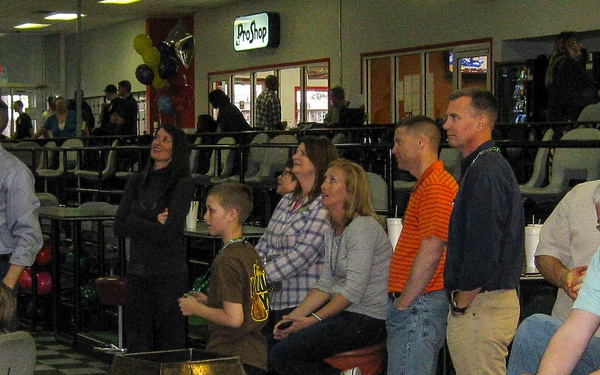 A group of adults and children are gathered at a bowling alley, many standing and watching something off-camera, likely ongoing bowling activity. The venue features a Pro Shop sign, bowling ball racks, scoring terminals, and checkered flooring typical of a bowling center. The atmosphere suggests a social event or party, with several people wearing Mardi Gras-style bead necklaces.