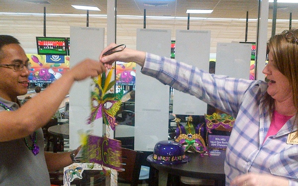 Two people are interacting at what appears to be a Mardi Gras themed event inside a bowling alley. A man wearing glasses and Mardi Gras beads and a woman in a plaid shirt with a name tag are holding up a colorful feathered Mardi Gras mask together. The setting includes Mardi Gras decorations, bowling lanes visible in the background, and a table with themed items.