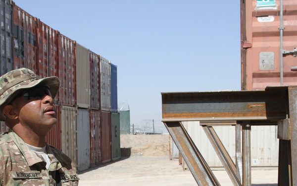A U.S. Army soldier in MultiCam/OCP camouflage uniform stands among stacked intermodal shipping containers in what appears to be a forward operating base or logistics yard in an arid environment. The soldier is looking upward and is armed with a rifle slung at his side. Patches and name tape are visible on the uniform, indicating active duty U.S. Army affiliation.