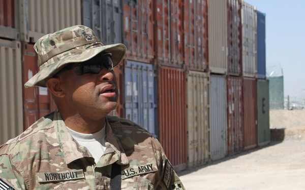 A U.S. Army soldier identified by name tape as 'Northcut' stands near stacked shipping containers in what appears to be a forward operating base or logistics area in an arid environment. The soldier is wearing MultiCam (OCP) camouflage uniform with a boonie hat, sunglasses, and is carrying a rifle. Visible patches indicate affiliation with the 4th Infantry Division.