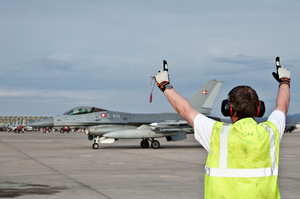 A ground crew marshal in a high-visibility yellow vest is directing a Danish Air Force F-16 Fighting Falcon (tail number E-611) on an airport tarmac. The aircraft displays the Danish flag roundel on its tail fin, identifying it as belonging to the Royal Danish Air Force. The marshal has both arms raised with thumbs up, indicating a standard marshalling signal.
