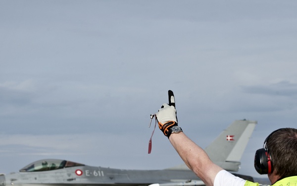 A ground crew marshal in a high-visibility yellow vest is directing a Danish Air Force F-16 Fighting Falcon (tail number E-611) on an airport tarmac. The aircraft displays the Danish flag roundel on its tail fin, identifying it as belonging to the Royal Danish Air Force. The marshal has both arms raised with thumbs up, indicating a standard marshalling signal.