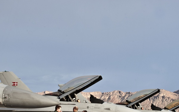 A line of F-16 Fighting Falcon jet aircraft are parked on a flight line apron, with canopies open and ground crew personnel conducting pre-flight or post-flight servicing. At least one aircraft displays a Danish flag roundel on its tail fin, identifying these as Royal Danish Air Force jets. The desert mountain terrain in the background is consistent with Nellis Air Force Base, Nevada, likely during a Red Flag exercise.