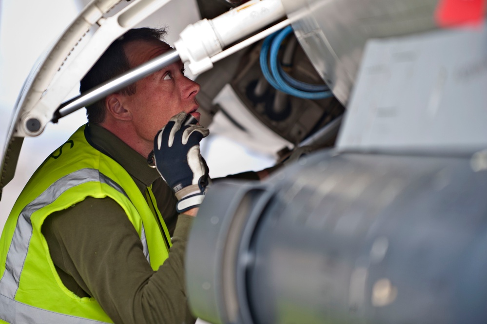 A maintenance technician wearing a high-visibility yellow safety vest and black work gloves is inspecting the underside of an aircraft, likely examining landing gear or an equipment bay. The worker is looking upward intently into the aircraft's mechanical compartment, with visible hydraulic lines, structural components, and what appears to be an external fuel tank or ordnance in the foreground. The image captures a close-up, detail-oriented maintenance inspection in an aviation environment.