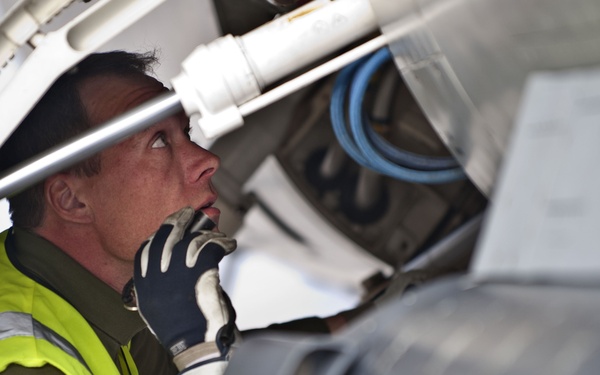 A maintenance technician wearing a high-visibility yellow safety vest and black work gloves is inspecting the underside of an aircraft, likely examining landing gear or an equipment bay. The worker is looking upward intently into the aircraft's mechanical compartment, with visible hydraulic lines, structural components, and what appears to be an external fuel tank or ordnance in the foreground. The image captures a close-up, detail-oriented maintenance inspection in an aviation environment.